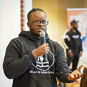 Reggie Howard stands with microphone in hand while speaking, wearing a black shirt that says "Black Men Heal"